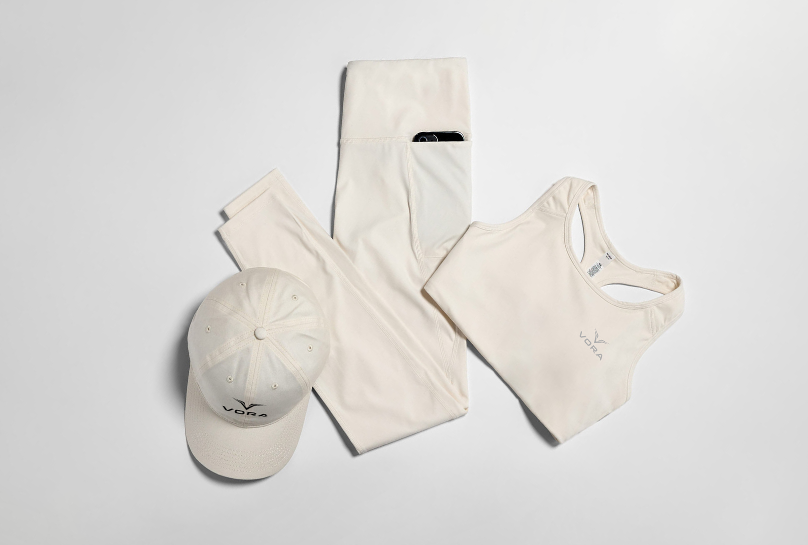Cream-colored activewear set includes a cap, leggings with a phone pocket, and a sports bra, all laid on a light background, conveying a minimalist and activewear vibe.