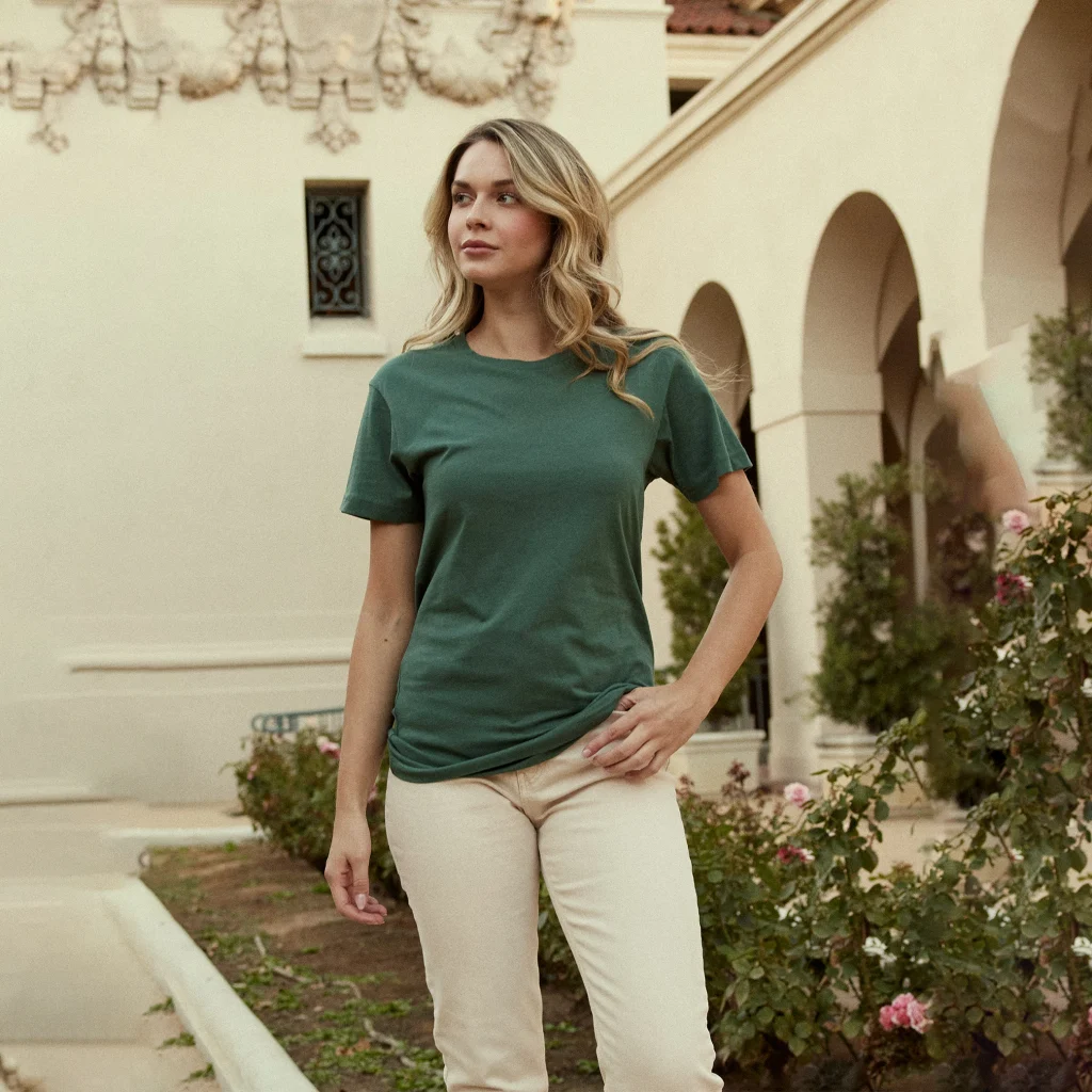 Woman wearing a forest green Cotton Heritage MC1040 unisex short sleeve T-shirt with cream pants outdoors.