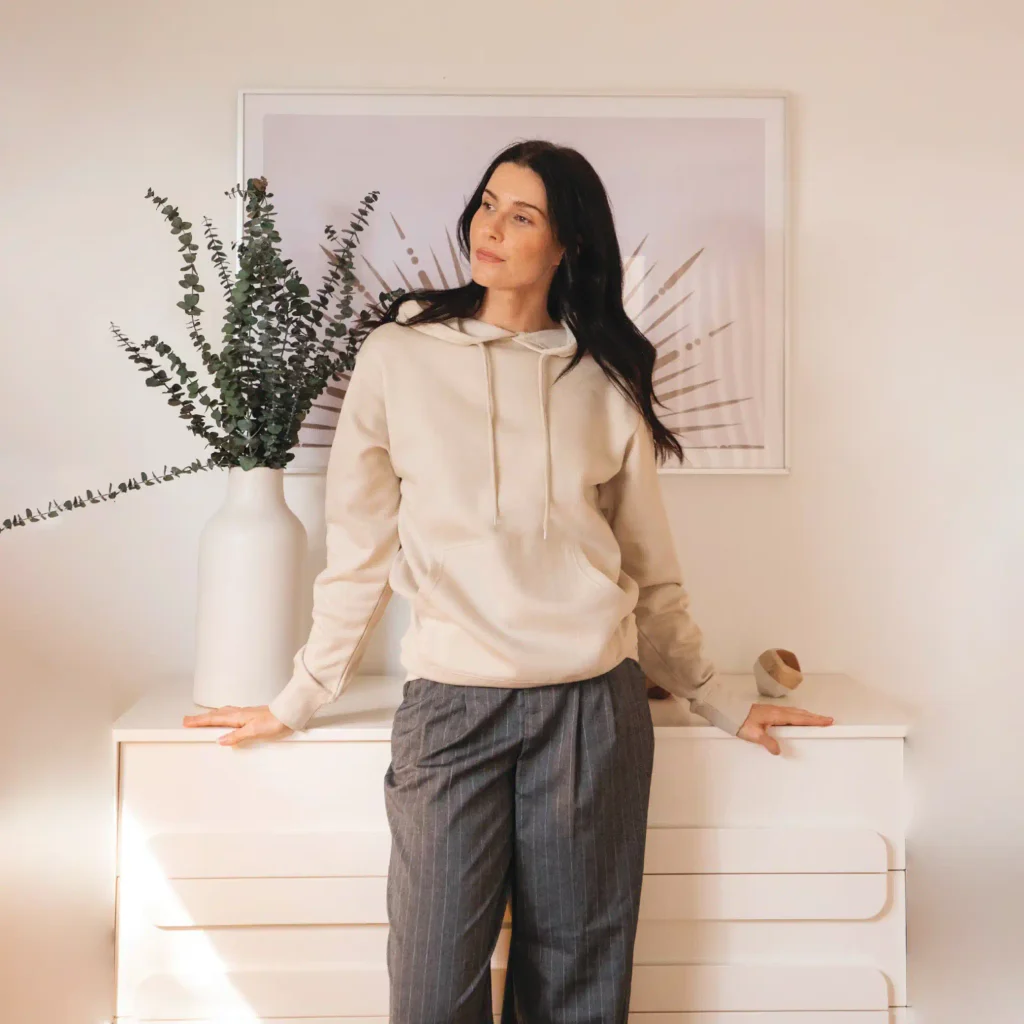 Woman wearing a bone Cotton Heritage M2500 lightweight pullover hoodie with dark gray trousers.